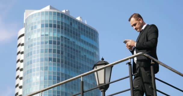 Man sms texting using app on smart phone in city. Handsome young businessman using smartphone smiling happy. Urban male professional commuting in his 30s against skyscraper and sky background - Footage, Video