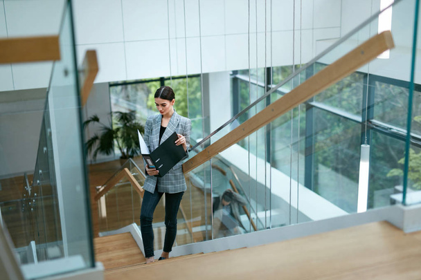 Business Woman Going To Work In Office. Portrait Of Successful Beautiful Female Worker In Stylish Clothes With Folder On Stairs. High Resolution - Фото, изображение