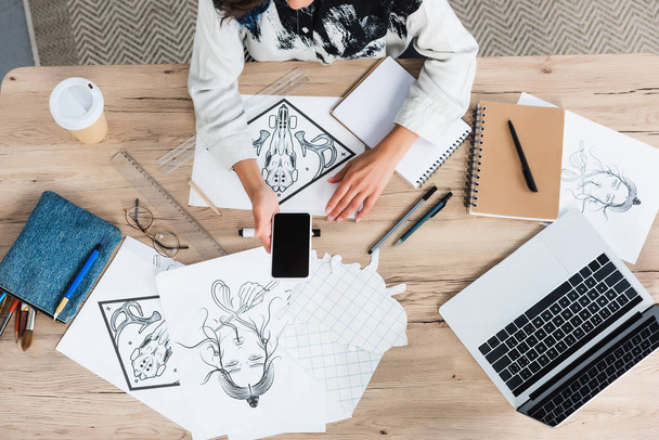 immagine ritagliata di designer donna che tiene smartphone con schermo bianco al tavolo di lavoro con dipinti e libri di testo
 - Foto, immagini