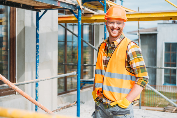 schöner lächelnder Bauarbeiter in reflektierender Weste und harter Mütze, der auf der Baustelle in die Kamera schaut - Foto, Bild