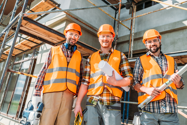 вид снизу группы счастливых оборудованных строителей, смотрящих на камеру на строительной площадке
 - Фото, изображение