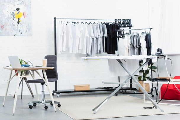 selective focus of t-shirts on hangers, table with laptop and ironing board in clothing design studio - Fotoğraf, Görsel