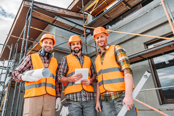 Blick von unten auf eine Gruppe lächelnder Bauarbeiter mit Blaupause und Tablet auf der Baustelle - Foto, Bild