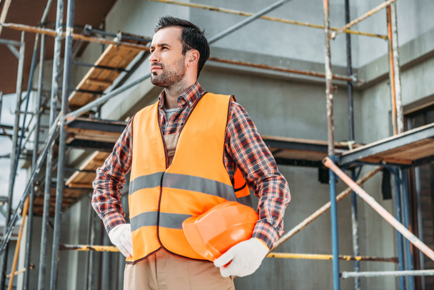 handsome builder in reflective vest holding helmet while standing in front of building house and looking away - Photo, Image
