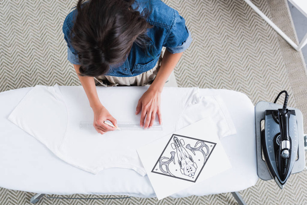 overhead view of female fashion designer measuring space for print on empty white t-shirt at ironing board - Foto, imagen