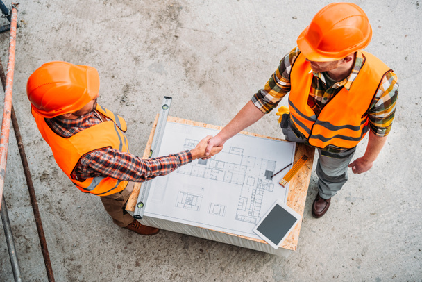 высокий угол обзора строителей, пожимающих руки над чертежом на строительной площадке
 - Фото, изображение
