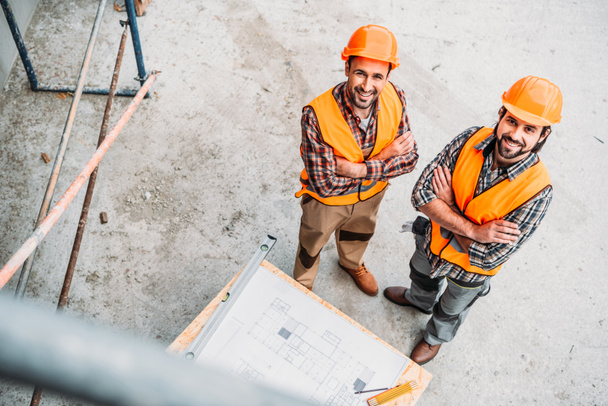 visão de alto ângulo de construtores sorridentes com planta em pé no local de construção e olhando para a câmera
 - Foto, Imagem