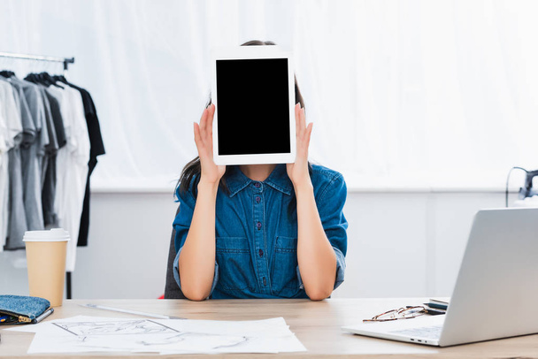 作業テーブルで空白の画面を持つデジタル タブレットで顔を覆っている若い女性  - 写真・画像