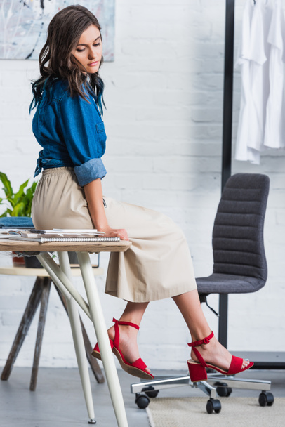 attractive female fashion designer sitting on table in clothing design studio - Photo, Image