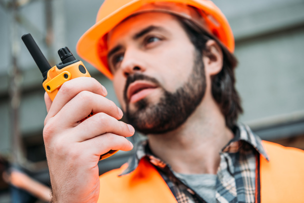 primer plano retrato del constructor en casco protector y chaleco usando walkie talkie en el sitio de construcción
 - Foto, Imagen