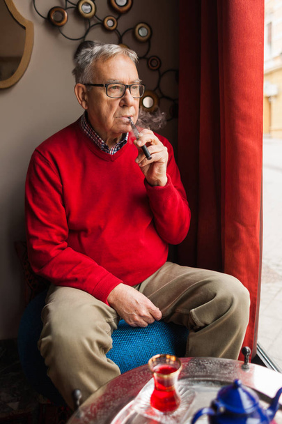 Senior man zitten in Cafe en elektronische sigaret roken.  - Foto, afbeelding