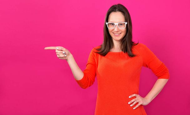 junge Frau, 25 Jahre alt, zeigt einen Finger, der auf einem rosa Hintergrund steht. schöne brünette Frau mit Brille. - Foto, Bild