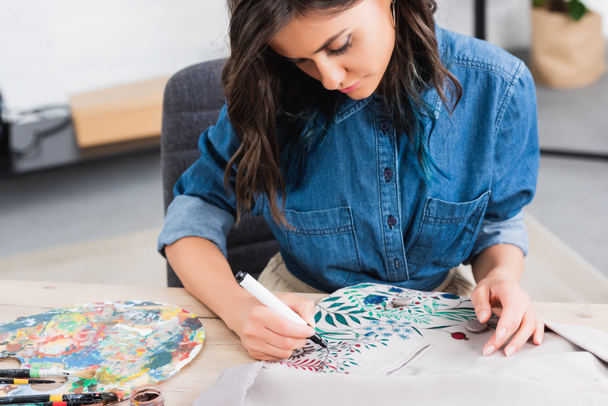 selective focus of female fashion designer painting on jacket at working table in clothing design studio - 写真・画像