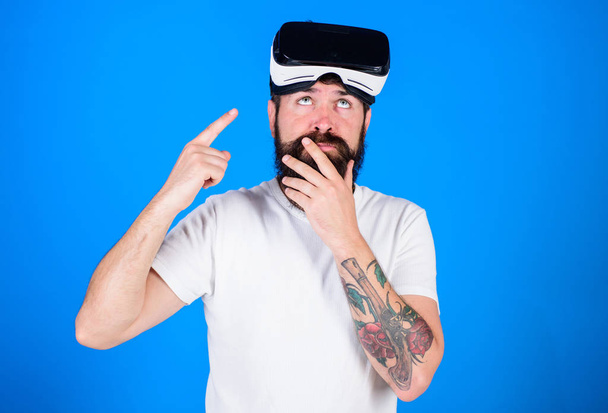 Man with beard and mustache with VR glasses, blue background. Guy with VR glasses or head mounted display. VR technology concept. Hipster on thoughtful face pointing at gadget with index finger - Foto, Bild