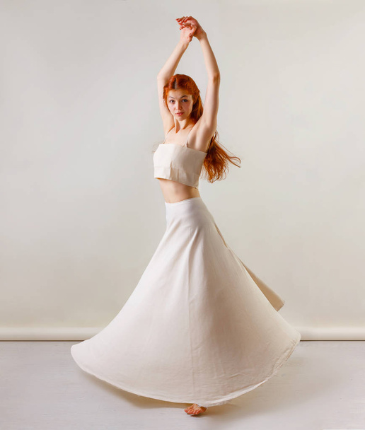 Tänzer posiert im Studio. junge und schöne rothaarige Mädchen in einem beigen langen Rock und Top tanzt und posiert im Studio. Kopierraum, grauer Hintergrund - Foto, Bild