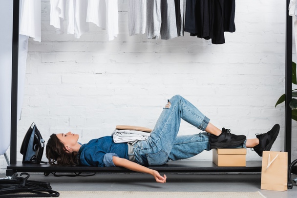 side view of female fashion designer laying with stack of white t-shirts and paper package in clothing design studio - Photo, Image