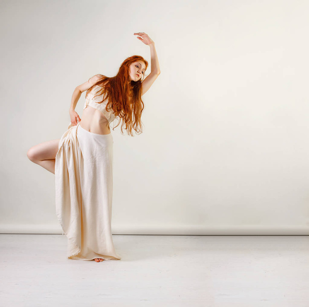 Dancer posing in studio. Young and beautiful redhead girl in a beige long skirt and top dances and poses in studio. Copy space - Photo, Image