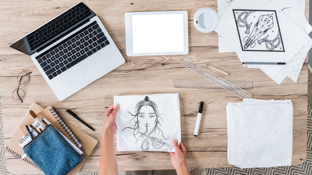 vista aerea di designer donna mettendo t-shirt bianca con stampa sul tavolo con dispositivi digitali e dipinti
  - Foto, immagini