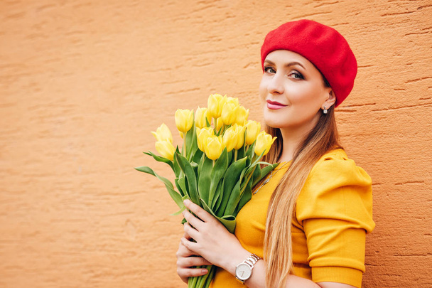Frühlingsporträt einer jungen schönen Frau, die gelbes Oberteil und rote Baskenmütze trägt, einen Strauß frischer Tulpenblumen in der Hand hält und gegen die orangefarbene Wand posiert - Foto, Bild