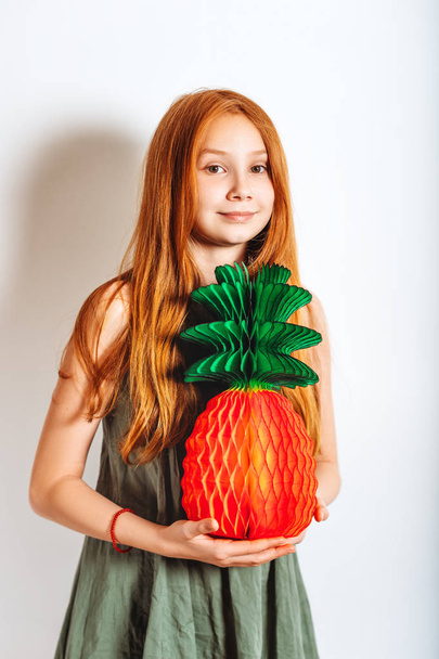 Studio shot of adorable red-haired preteen kid girl, wearing khaki dress, playing with paper pineapple - Photo, Image