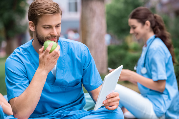 ハンサムな白人医学生リンゴを食べるとタブレットを見て - 写真・画像