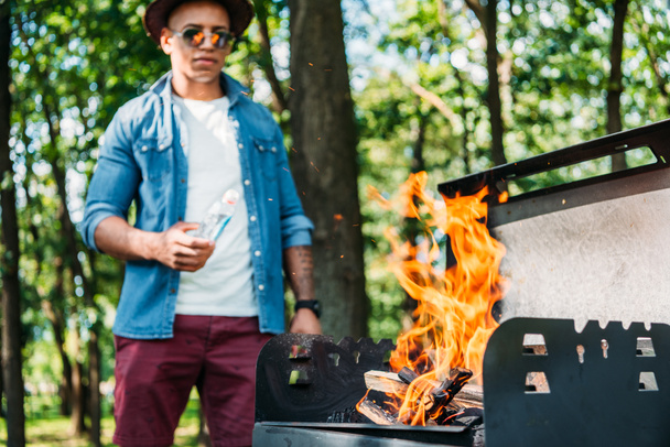 selective focus of african american man in sunglasses and hat setting fire on grill in park - Photo, Image