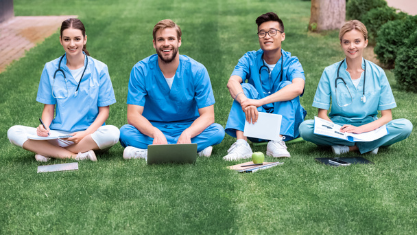 мультикультурные студенты-медики сидят на траве и смотрят в камеру
 - Фото, изображение