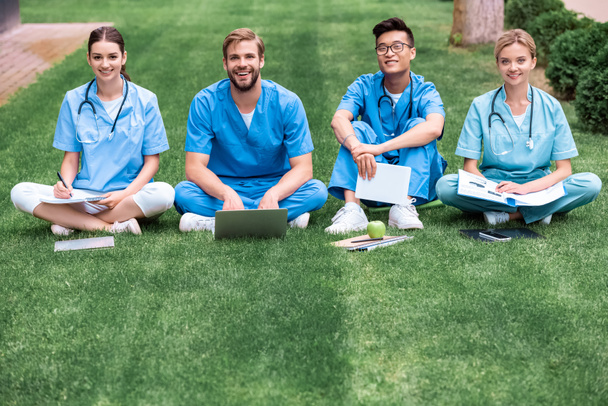 Lächelnde multikulturelle Medizinstudenten, die auf Gras sitzen und in die Kamera schauen - Foto, Bild