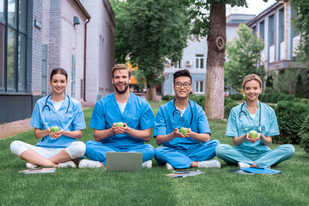 happy multicultural medical students sitting on grass and holding apples - Foto, Imagem