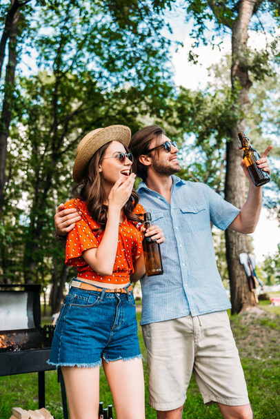 cheerful couple in sunglasses with beer looking away in park - Fotó, kép