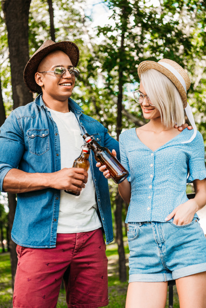 portrait of multiracial couple clinking bottles of beer in summer park - Фото, изображение