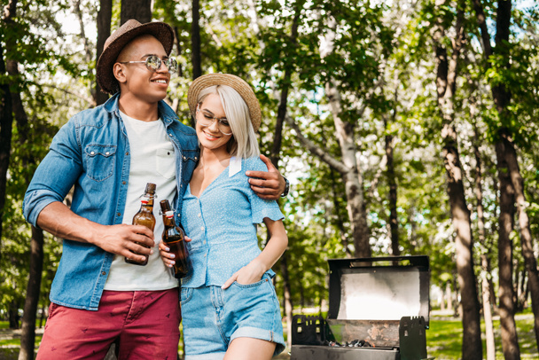 portrait of multiracial couple with beer standing near grill in summer park - Фото, зображення