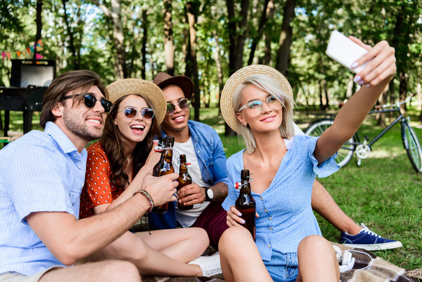 smiling multicultural friends with beer taking selfie on smartphone while resting in summer park - Foto, afbeelding