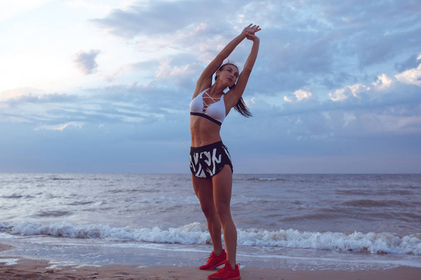 the girl does a warm-up before doing sports early in the morning on the seashore. A girl of athletic build with a dry muscle mass. - Foto, immagini
