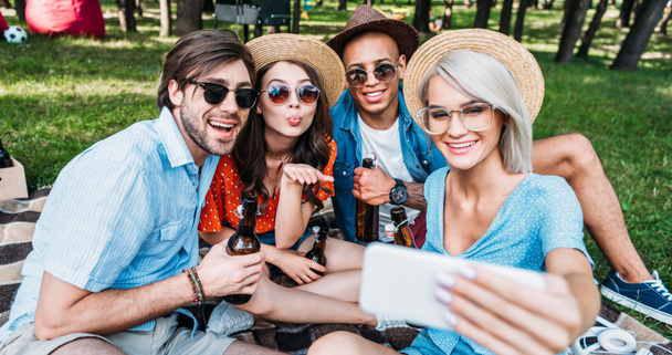 amigos multiculturais sorridentes com cerveja tomando selfie no smartphone enquanto descansa no parque de verão
 - Foto, Imagem