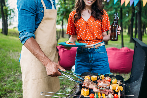 vue partielle de la femme avec de la bière regardant le copain afro-américain cuisiner des aliments sur le gril pendant le barbecue
 - Photo, image