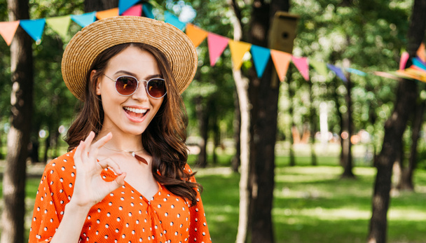 portrait of happy young woman in hat and sunglasses showing ok sign in park - Photo, Image