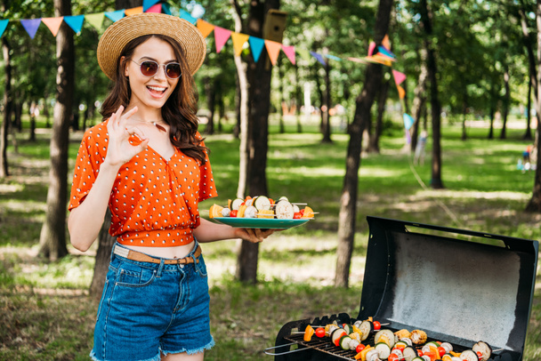 portrait of happy woman in hat and sunglasses with grilled vegetables on plate showing ok sign in park - Foto, Bild