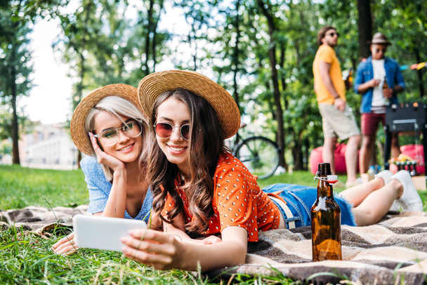 selective focus of smiling friends taking selfie on smartphone while resting on blanket during bbq in park - Foto, imagen