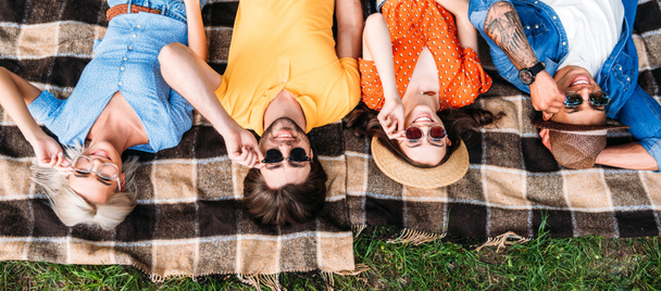 overhead view of interracial friends in sunglasses resting on blanket in park - Foto, afbeelding