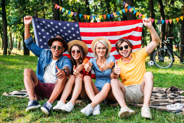 glückliche multirassische Freunde mit amerikanischer Flagge, die Daumen hoch im Sommerpark zeigt - Foto, Bild