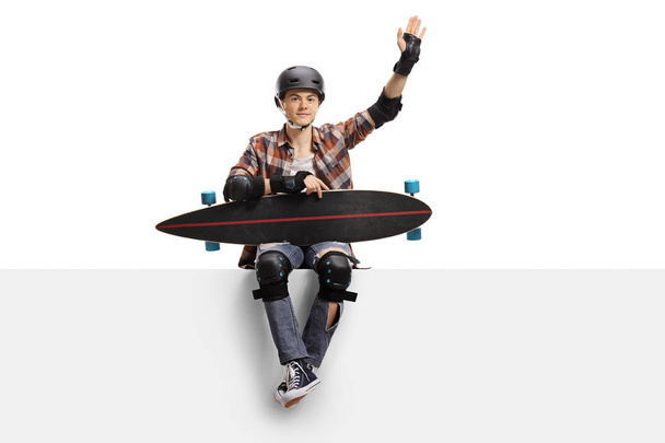 Teenage skater with a longboard and protective equipment sitting on a panel and waving isolated on white background - Fotografie, Obrázek