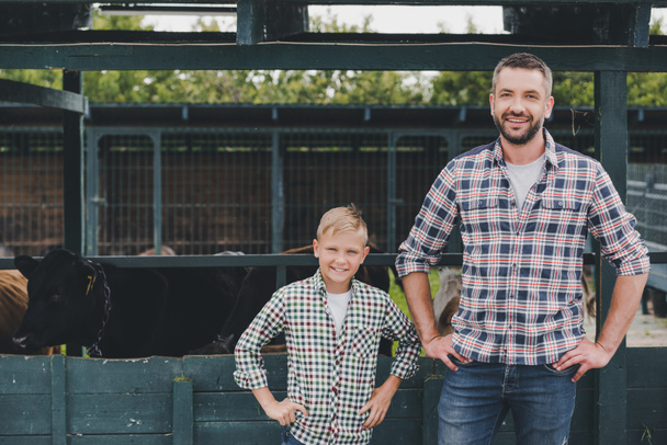 heureux père et fils debout avec les mains sur la taille et souriant à la caméra à la ferme
 - Photo, image