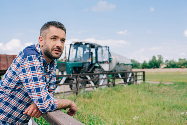 pensive middle aged farmer leaning at railing and looking away at farm  - Фото, изображение