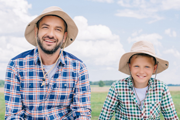 happy father and son in checkered shirts and panama hats smiling at camera outdoors - 写真・画像