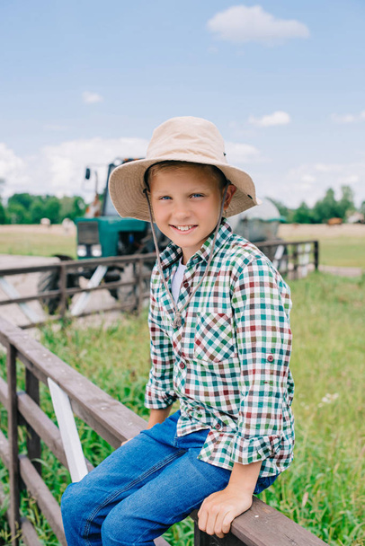 cheerful boy in panama hat sitting on fence and smiling at camera on farm  - Фото, зображення