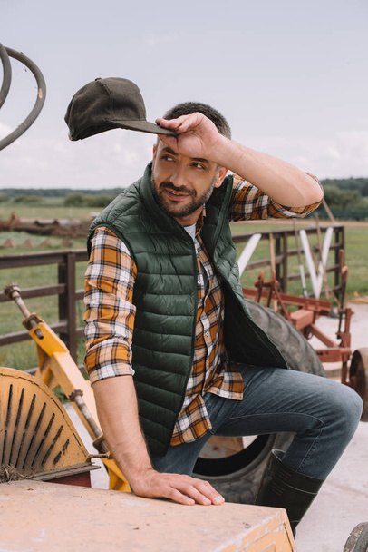 handsome male farmer holding cap and looking away while working at ranch - 写真・画像