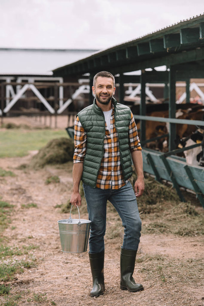 handsome male farmer holding bucket and smiling at camera at cowshed - Foto, Bild
