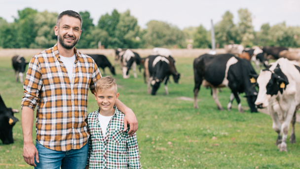 ευτυχής πατέρας και γιος χαμογελά στη φωτογραφική μηχανή, ενώ στέκεται κοντά σε βόσκηση βοοειδών στο αγρόκτημα  - Φωτογραφία, εικόνα
