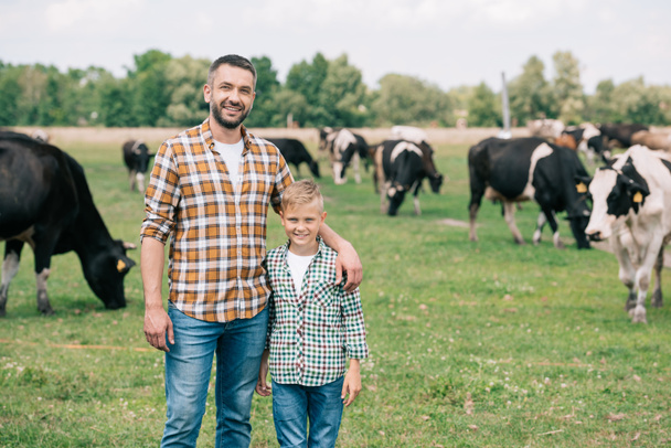 father and son smiling at camera while standing near grazing cattle at farm  - Photo, image